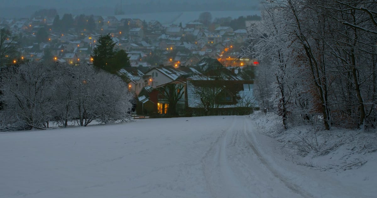 Verschneite Straße am Ortsrand in der Dämmerung, dahinter beleuchtete Häuser in einem Tal.